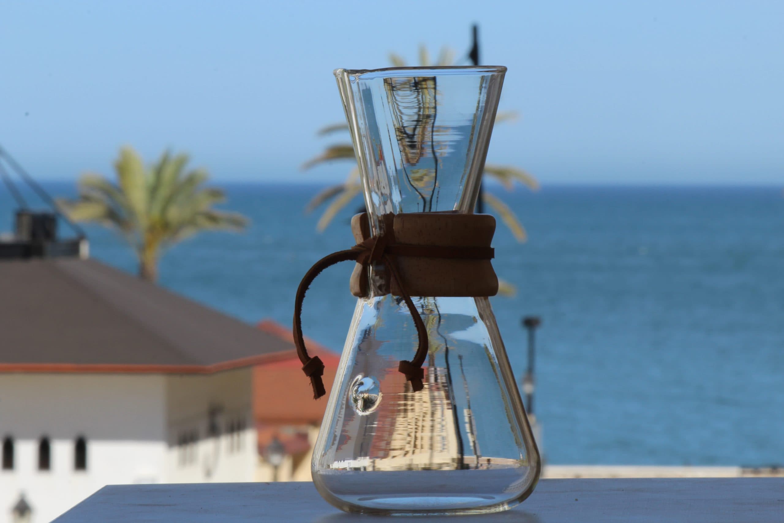 Chemex de cristal con collar de madera sobre una mesa, con el mar Mediterráneo y palmeras desenfocadas al fondo