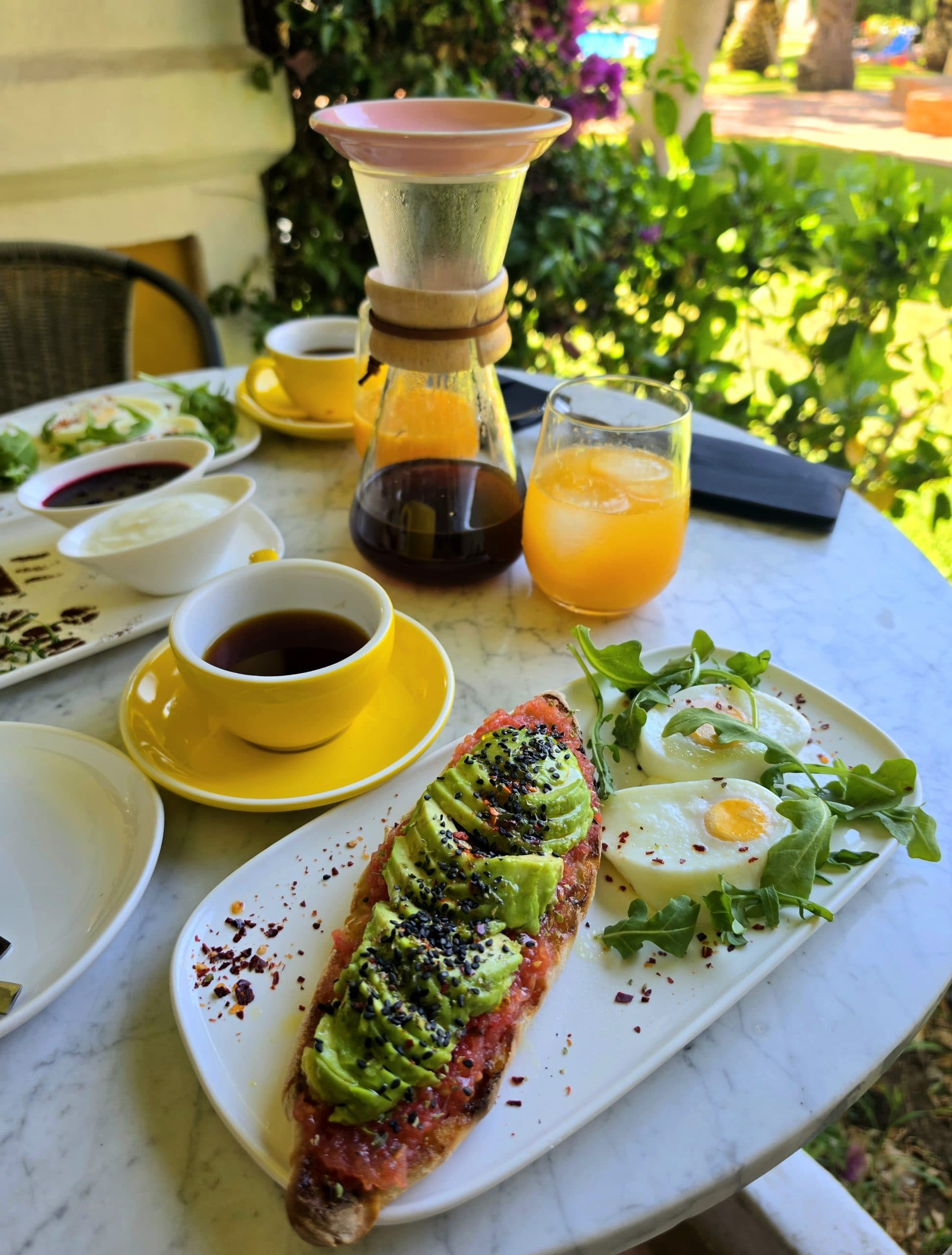 Tosta de aguacate con huevo y brotes verdes sobre mesa de mármol, con taza amarilla de café, zumo de naranja y una Chemex al fondo