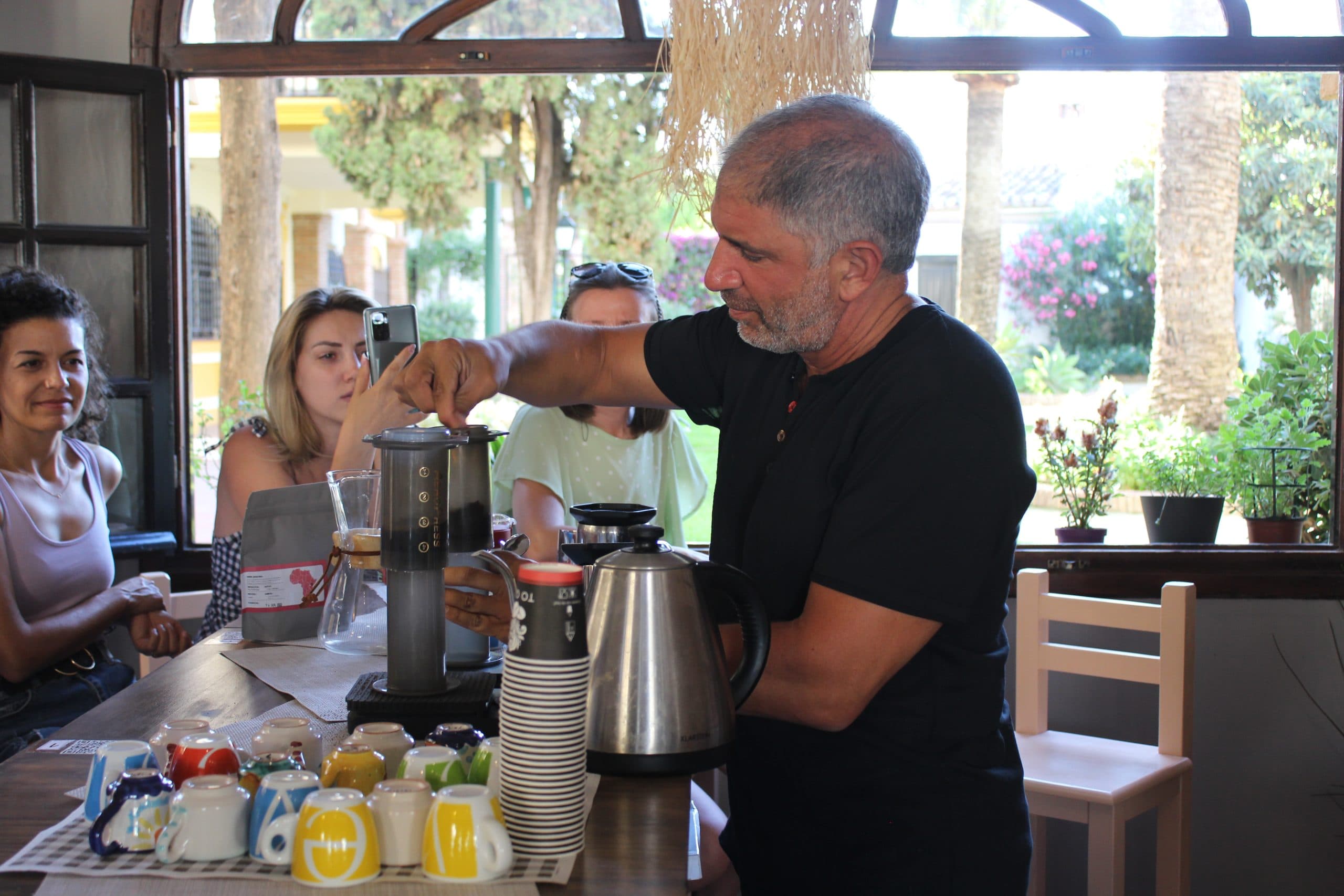 Un barista prepara café con AeroPress ante un grupo de asistentes sentados a la mesa, con hervidor de cuello de cisne, café de origen y tazas de colores alineadas