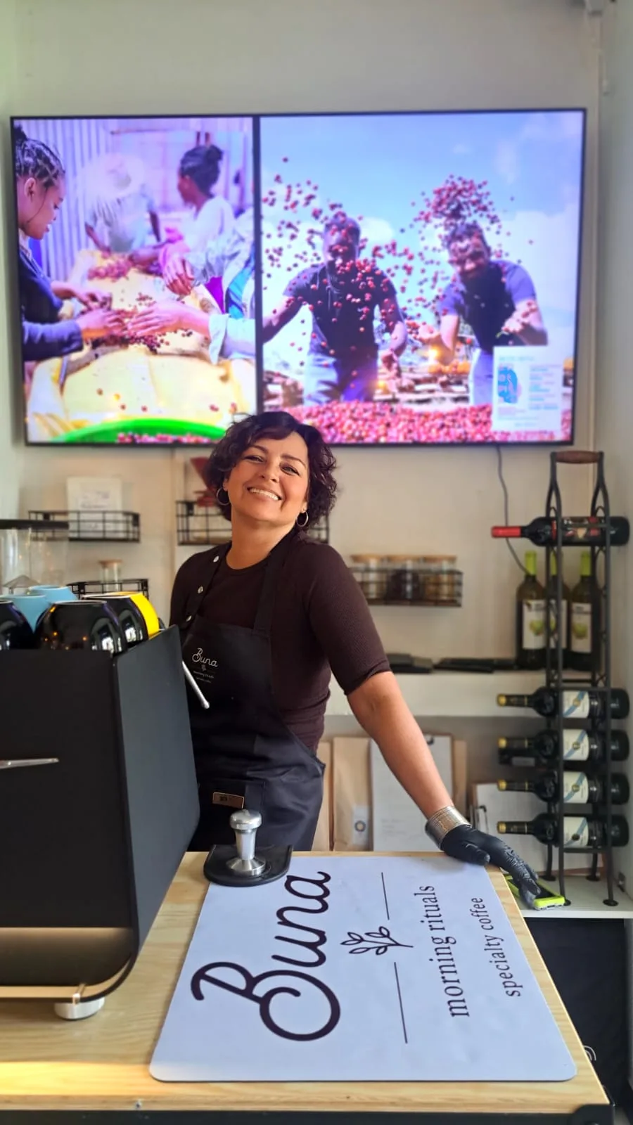 Buna barista smiling behind the counter, next to the espresso machine and the tray with the Buna logo