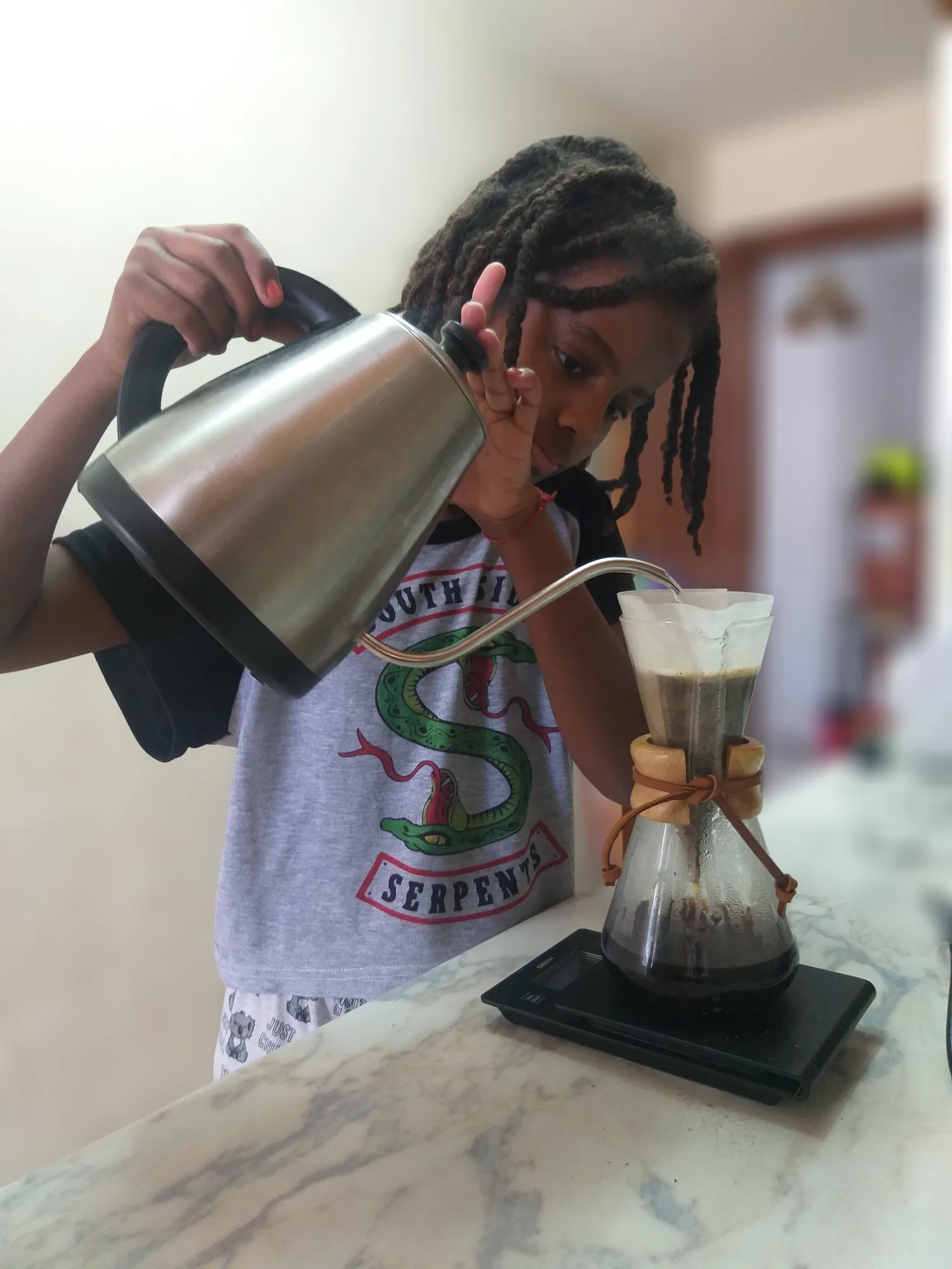 A girl pours water from a gooseneck kettle over a Chemex resting on a scale, on a marble kitchen counter