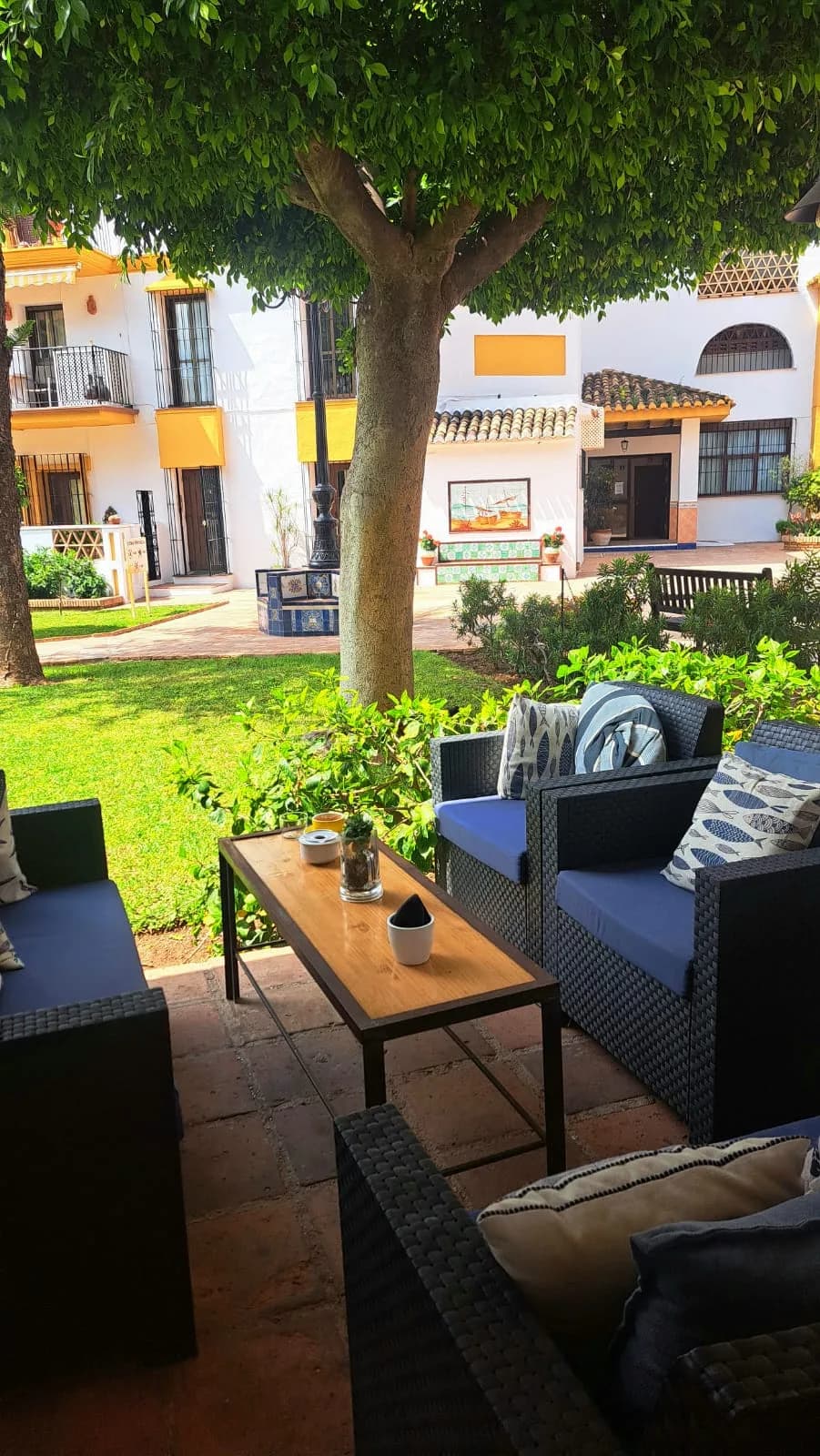 Sofás de exterior con cojines azules alrededor de una mesa baja, a la sombra de un árbol, frente al césped y los edificios blancos del complejo