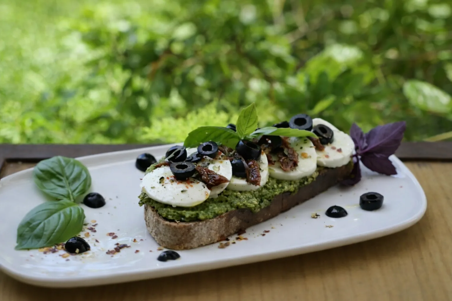 Sourdough toast with pesto, mozzarella, sun-dried tomato, black olives and basil, on a white plate on a wooden garden table