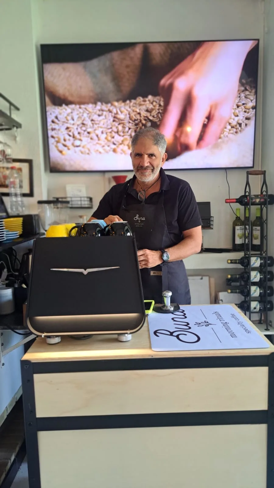 A Buna team member in a house apron smiles behind the counter next to the tabletop coffee roaster, with a screen showing coffee beans behind