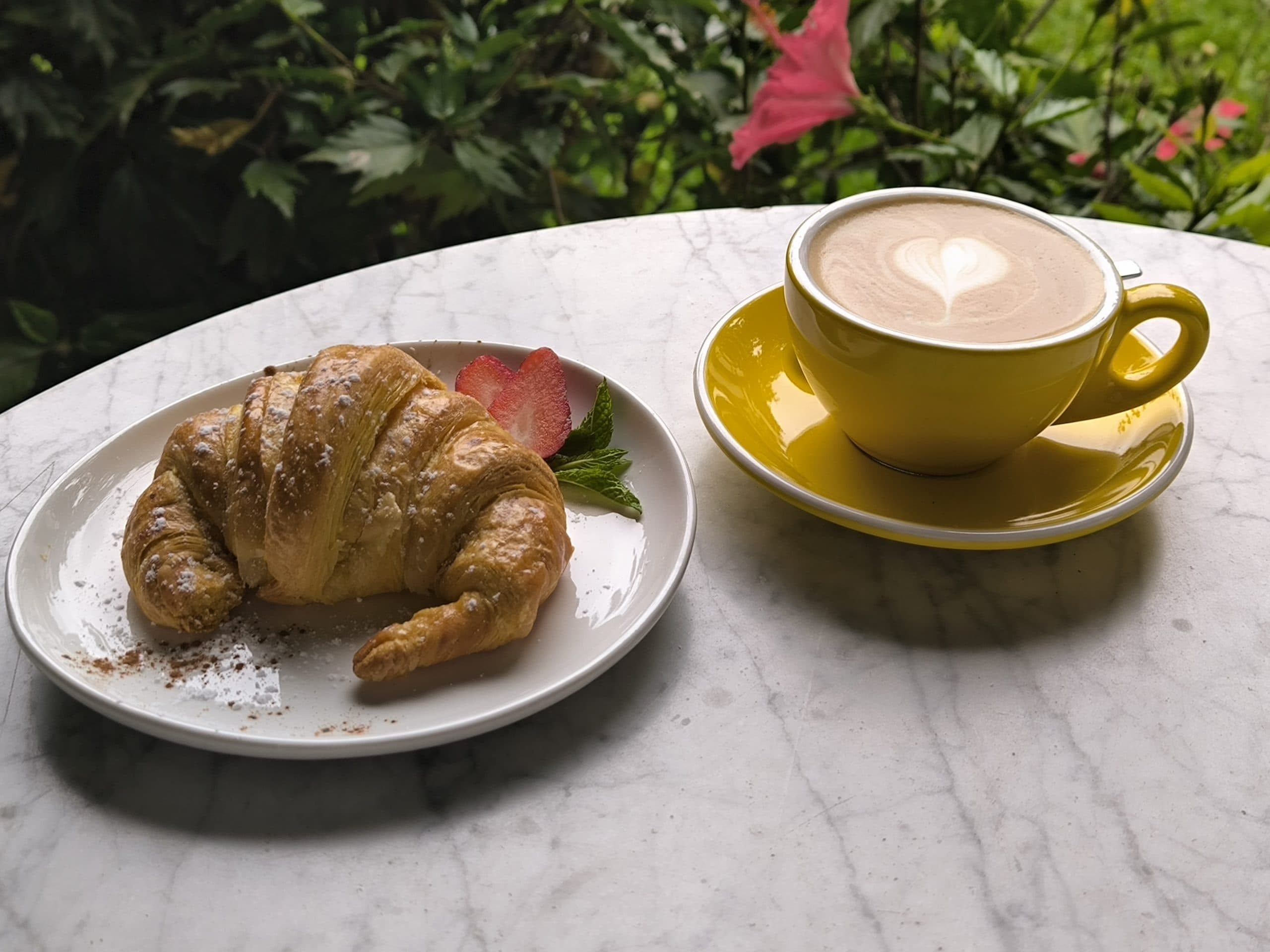 Capuchino con arte latte en taza amarilla junto a un croissant sobre mármol, con la vegetación del jardín al fondo
