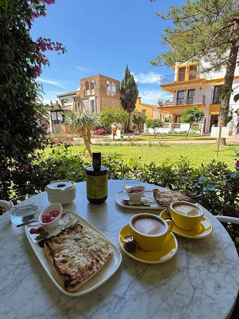 Mesa de mármol servida en el jardín de Aidan's Bar, con tazas amarillas y el edificio entre las palmeras al fondo
